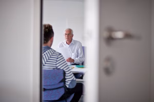 Older man with white hair presenting a document to a younger colleague across a table in an office meeting room]