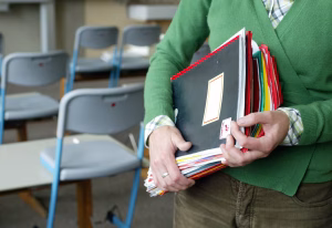 Person in a green sweater holds a stack of folders and notebooks in a classroom setting, with plaid shirt cuffs visible.