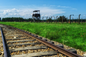 Railroad tracks run through a grassy field toward a tall wooden lookout tower and a barbed-wire perimeter fence under a blue sky.