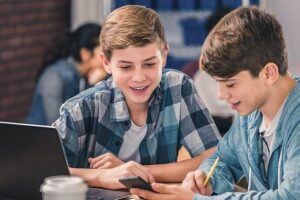Two teenage boys study together at a table with a laptop, phone, and notebook in a classroom setting.