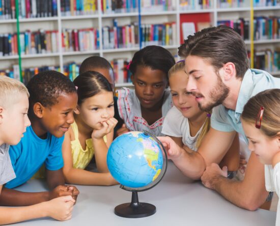 Teacher and diverse group of children gathered around a globe in a library, examining maps together.