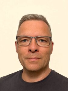 Head-and-shoulders portrait of a man with gray hair and glasses wearing a dark T-shirt against a light background, neutral expression