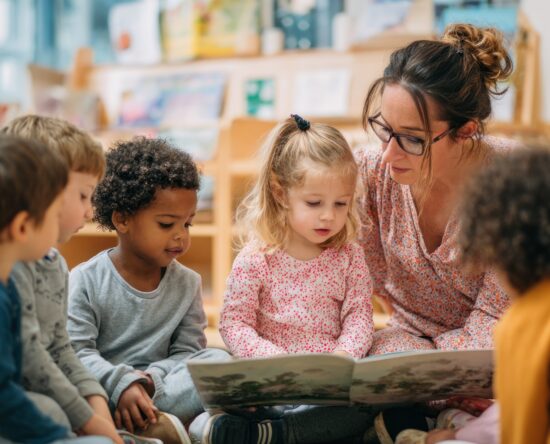Teacher reads aloud to a diverse group of children gathered around a big picture book in a library or classroom setting.