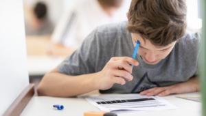 Boy focused on a worksheet at a white desk, holding a blue pen over the page with markers nearby.