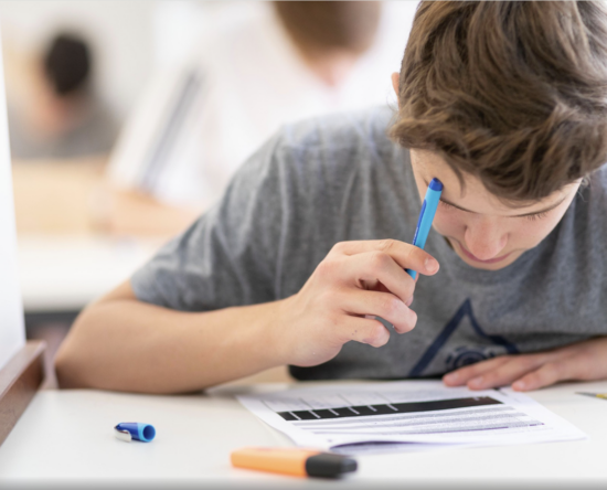 Boy focused on a worksheet at a white desk, holding a blue pen over the page with markers nearby.