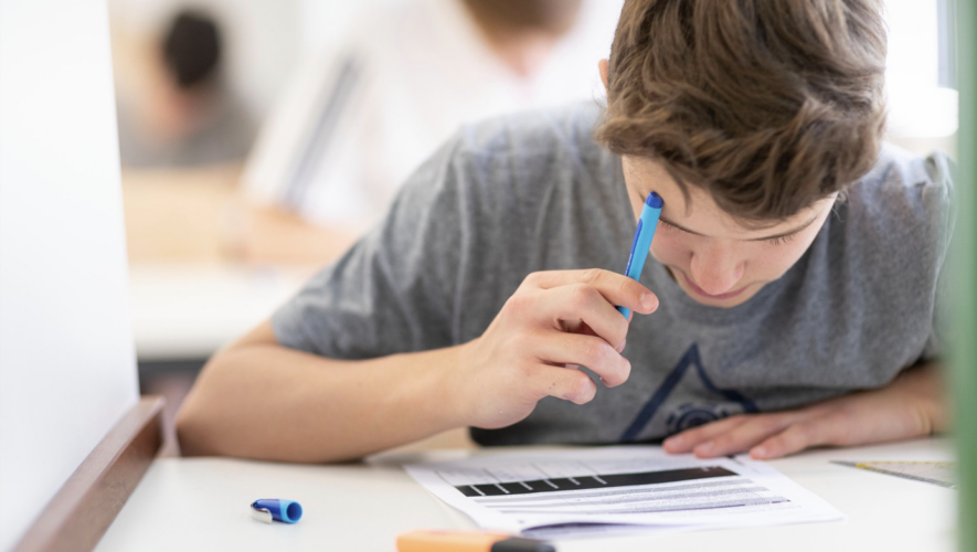 Boy focused on a worksheet at a white desk, holding a blue pen over the page with markers nearby.