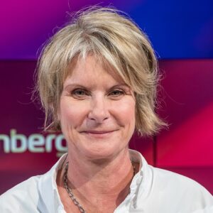 Portrait of a smiling middle-aged woman with short blonde hair, wearing a white collared shirt and a silver chain necklace, against a purple-pink studio backdrop