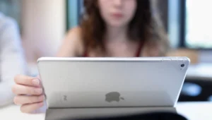 Person holding a silver tablet (back view) indoors, with a blurred person in the background.