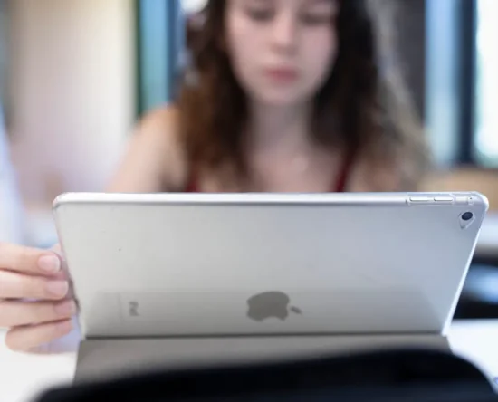 Person holding a silver tablet (back view) indoors, with a blurred person in the background.