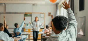 Teacher standing at the front of a classroom as students raise their hands to answer a question in a bright, engaged class.