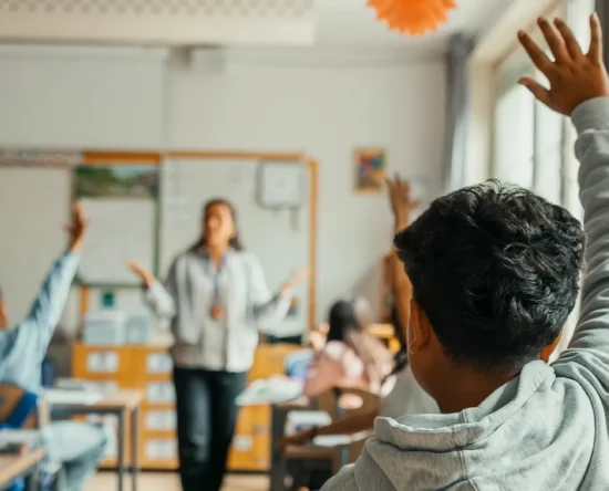 Teacher standing at the front of a classroom as students raise their hands to answer a question in a bright, engaged class.