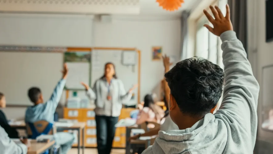Teacher standing at the front of a classroom as students raise their hands to answer a question in a bright, engaged class.