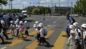 Group of young children in white hats crossing a yellow crosswalk under adult supervision in an urban street.