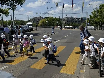 Group of young children in white hats crossing a yellow crosswalk under adult supervision in an urban street.
