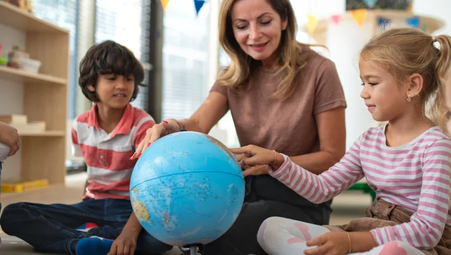 Teacher guiding a group of children as they explore a globe in a bright classroom.