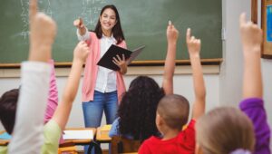 Teacher at a chalkboard holds a notebook while students raise their hands to answer in a classroom scene