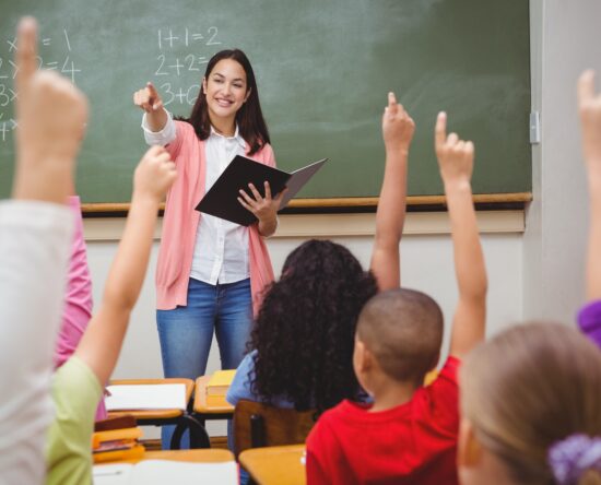 Teacher at a chalkboard holds a notebook while students raise their hands to answer in a classroom scene