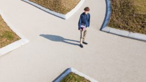 Overhead view of a young man in a blue hoodie standing on a curved concrete path, holding a binder or laptop case.