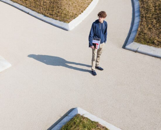 Overhead view of a young man in a blue hoodie standing on a curved concrete path, holding a binder or laptop case.