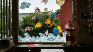 Colorful paper sea creatures stuck to a large classroom window, with a red-and-white lighthouse model on the desk inside.