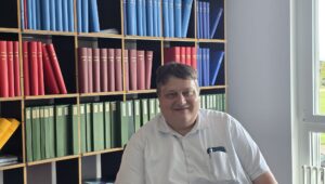 Smiling man in a white shirt seated in front of wooden shelves filled with blue, red, and green binders in an office.