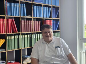 Smiling man in a white shirt seated in front of wooden shelves filled with blue, red, and green binders in an office.