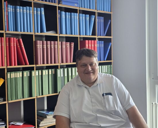 Smiling man in a white shirt seated in front of wooden shelves filled with blue, red, and green binders in an office.