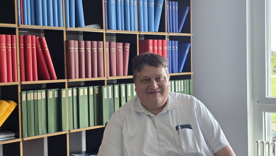Smiling man in a white shirt seated in front of wooden shelves filled with blue, red, and green binders in an office.