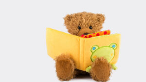 Brown teddy bear sitting and reading a yellow book with a frog illustration on the cover.