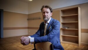 Portrait of a bearded man in a blue blazer sitting on a chair in an empty room with wooden floors and shelves in the background, looking at the camera.