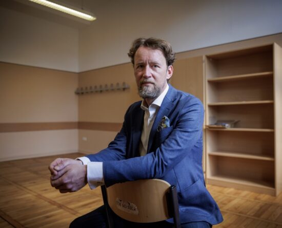 Portrait of a bearded man in a blue blazer sitting on a chair in an empty room with wooden floors and shelves in the background, looking at the camera.