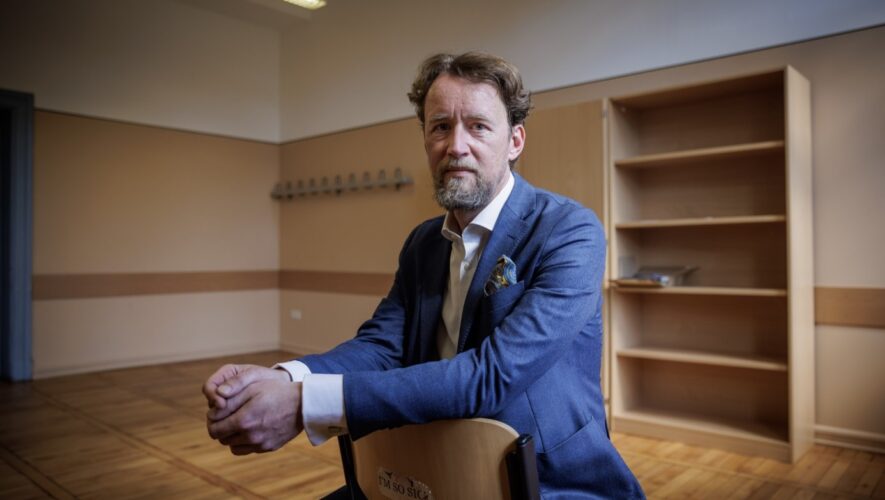 Portrait of a bearded man in a blue blazer sitting on a chair in an empty room with wooden floors and shelves in the background, looking at the camera.