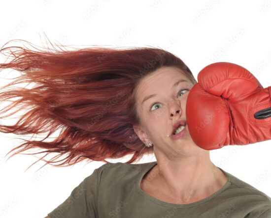Woman with red hair is punched in the face by a large red boxing glove, hair flying and eyes widened in surprise.
