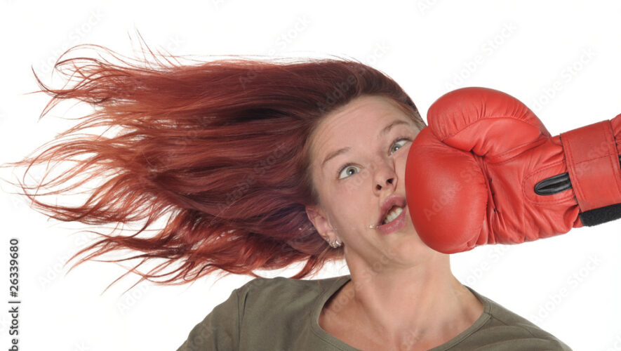 Woman with red hair is punched in the face by a large red boxing glove, hair flying and eyes widened in surprise.