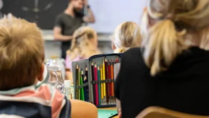 Students in a classroom with a teacher at the front; a pencil case full of colored pencils sits on a desk in the foreground.