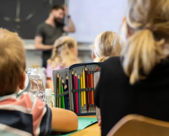 Students in a classroom with a teacher at the front; a pencil case full of colored pencils sits on a desk in the foreground.