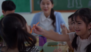 Girl with braided hair smiles as she hands an orange utensil to a classmate at a lunch table, with a teacher in the background.]} }} Wait formatting: need proper JSON array of multiple strings. Let's craft again.
