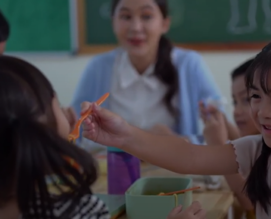 Girl with braided hair smiles as she hands an orange utensil to a classmate at a lunch table, with a teacher in the background.]} }} Wait formatting: need proper JSON array of multiple strings. Let's craft again.