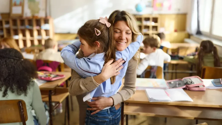 Teacher hugs a smiling student in a bustling classroom environment.