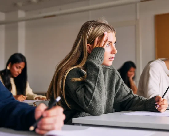 Group of students in a classroom taking notes; a young woman in a gray sweater holds her head with a worried expression as others write nearby.