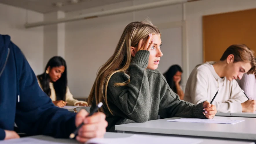 Group of students in a classroom taking notes; a young woman in a gray sweater holds her head with a worried expression as others write nearby.