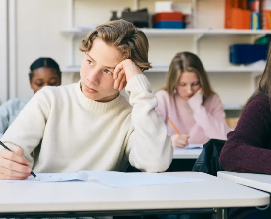 Students sit at desks in a classroom, looking bored or tired while writing on papers in front of them.