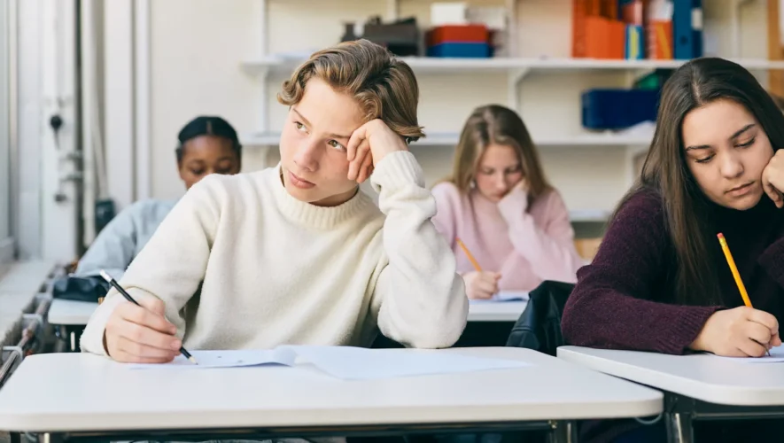Students sit at desks in a classroom, looking bored or tired while writing on papers in front of them.