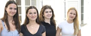 Four young women stand together outside a building, smiling at the camera, wearing casual summer outfits.
