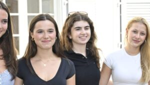 Four young women stand together outside a building, smiling at the camera, wearing casual summer outfits.