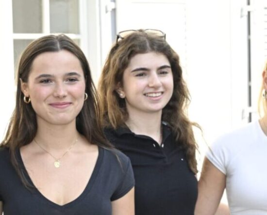 Four young women stand together outside a building, smiling at the camera, wearing casual summer outfits.