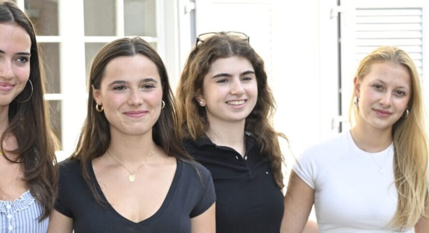 Four young women stand together outside a building, smiling at the camera, wearing casual summer outfits.
