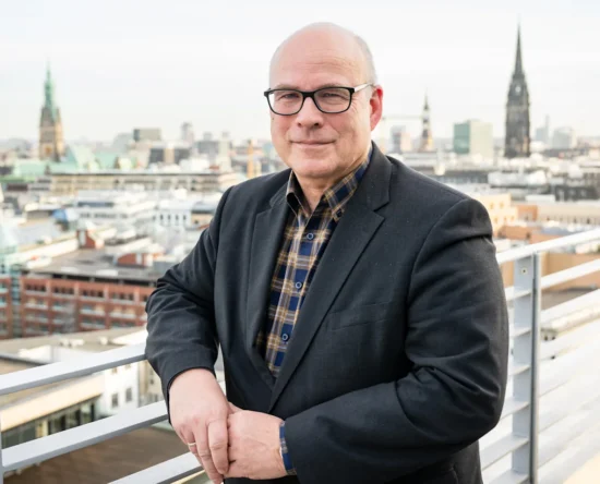 Portrait of a middle-aged man in a dark blazer and plaid shirt standing on a rooftop balcony with a city skyline behind him, smiling. This is a professional portrait with an urban backdrop.