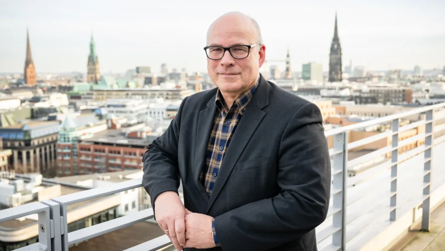 Portrait of a middle-aged man in a dark blazer and plaid shirt standing on a rooftop balcony with a city skyline behind him, smiling. This is a professional portrait with an urban backdrop.